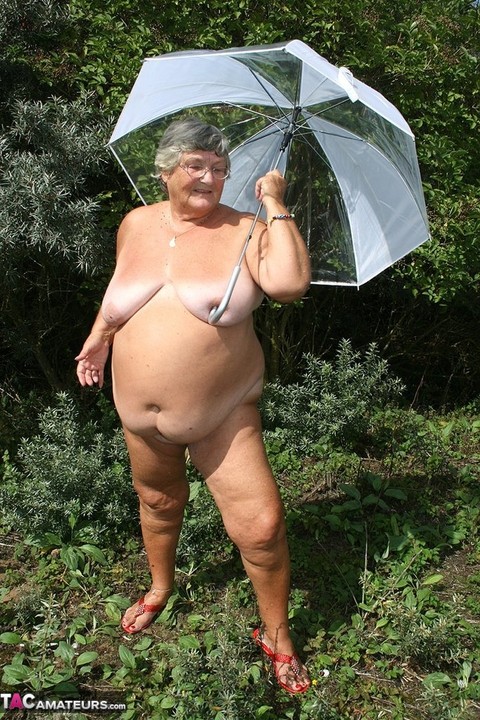 Obese oma Grandma Libby holds an umbrella while posing naked by fir trees | Фото 15