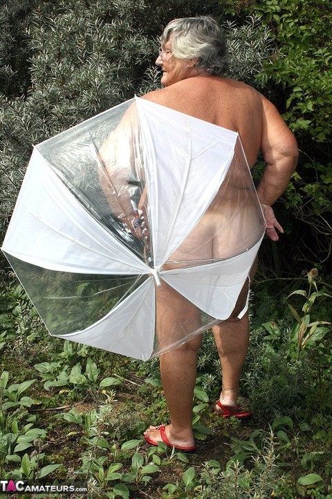 Obese oma Grandma Libby holds an umbrella while posing naked by fir trees | Фото 2