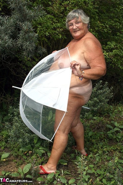 Obese oma Grandma Libby holds an umbrella while posing naked by fir trees | Фото 3