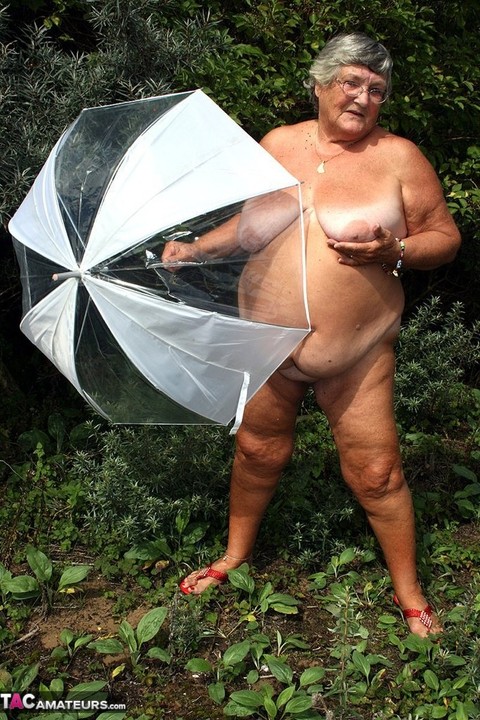 Obese oma Grandma Libby holds an umbrella while posing naked by fir trees | Фото 4