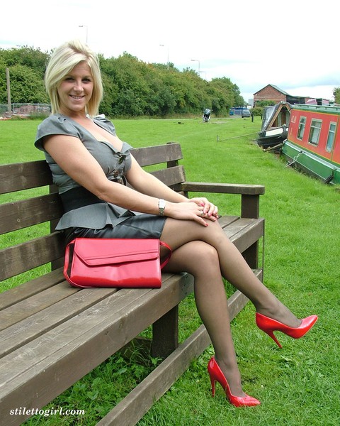 Clothed female with blonde hair shows off her red pumps in nylons on a bench | Фото 1